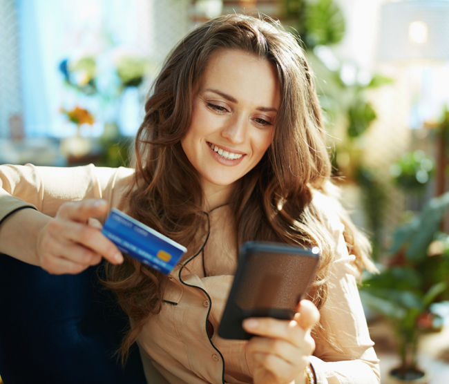 Green Home. happy trendy middle aged woman with long wavy hair with credit card making online shopping on e-commerce website in the modern house in sunny day.