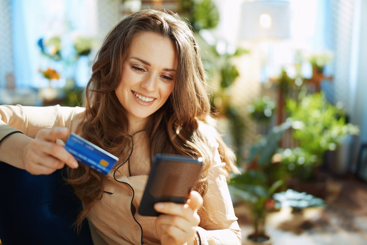 Green Home. happy trendy middle aged woman with long wavy hair with credit card making online shopping on e-commerce website in the modern house in sunny day.