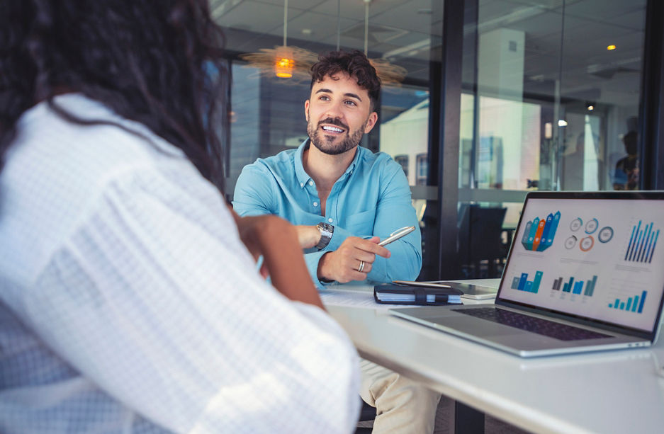 Business colleagues having a conversation. They are discussing a charts and graphs on a laptop computer. They are both young business people casually dressed in an office. Could be an interview or consultant working with a client.