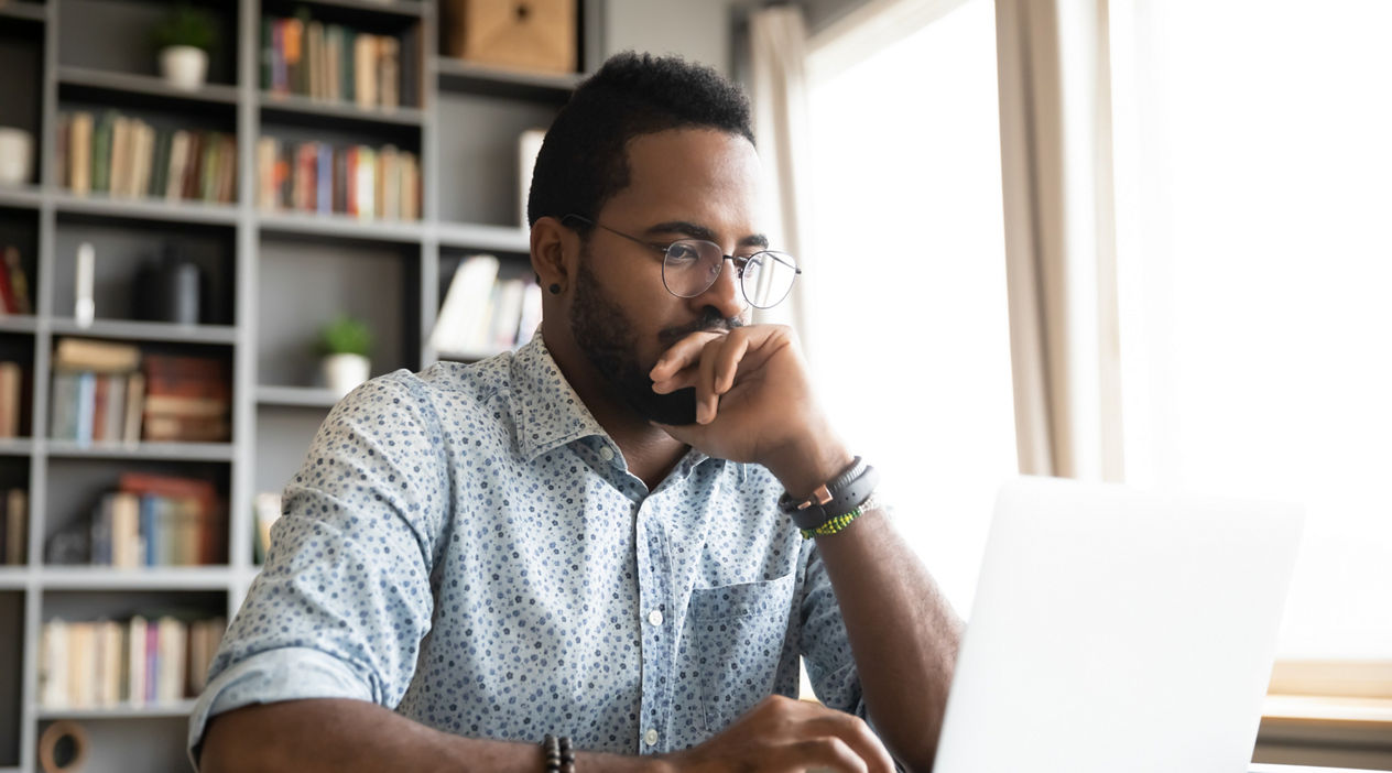 Focused concentrated young african businessman sit at desk look at laptop, serious afro american male professional analyst working online on computer data watch webinar thinking of problem solution