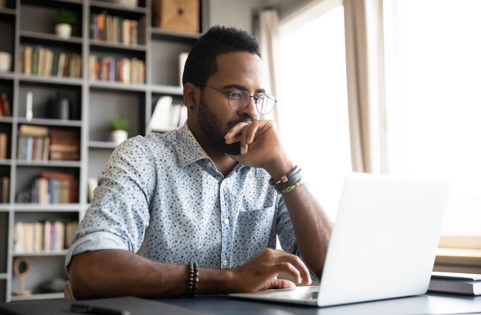 Focused concentrated young african businessman sit at desk look at laptop, serious afro american male professional analyst working online on computer data watch webinar thinking of problem solution