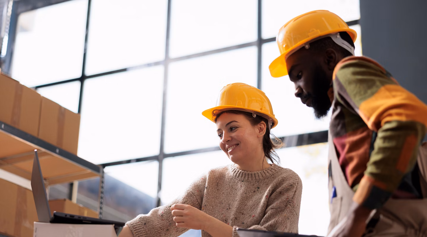 Diverse coworkers wearing industrail overall and helmet, while checking online orders on laptop computer in storage room. Stockroom team analyzing delivery details on digital device in warehouse