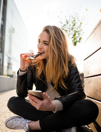 Wide angle photograph of teenager eating chocolate on phone outddoor in street sitting on bench