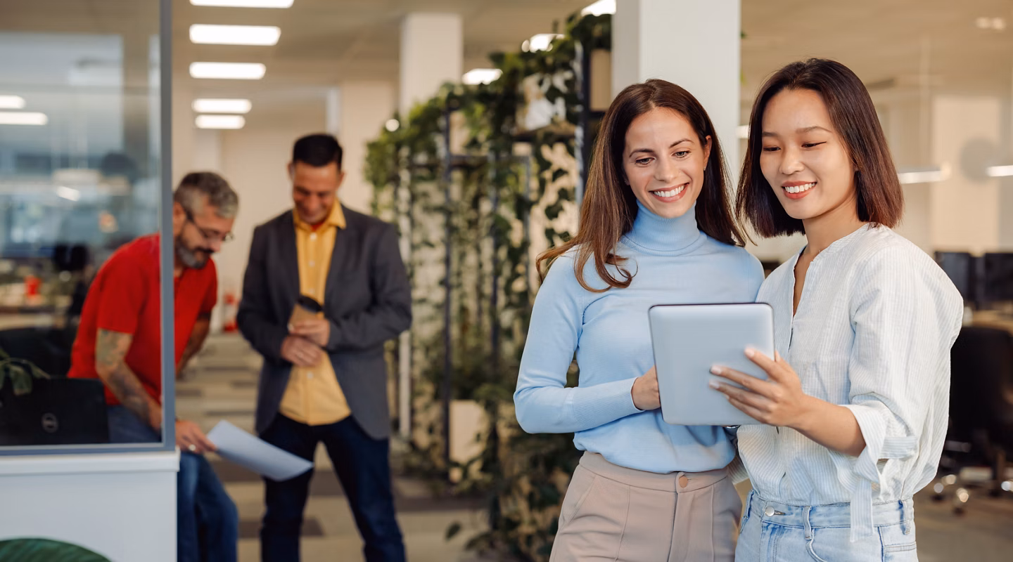 Portrait of a two diverse female business colleagues working together on a digital tablet. Multiracial creative executives meeting in an office and smiling. Male colleagues in the back. Copy space.