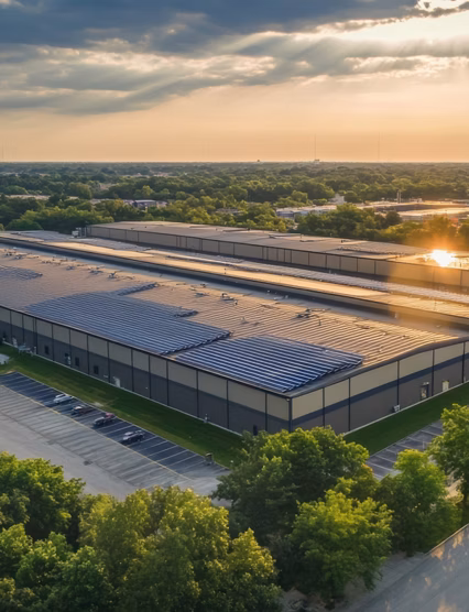 Aerial perspective showcases a sizable warehouse complex with solar panels, bordered by greenery and grass, illuminated by the warm light of sunset