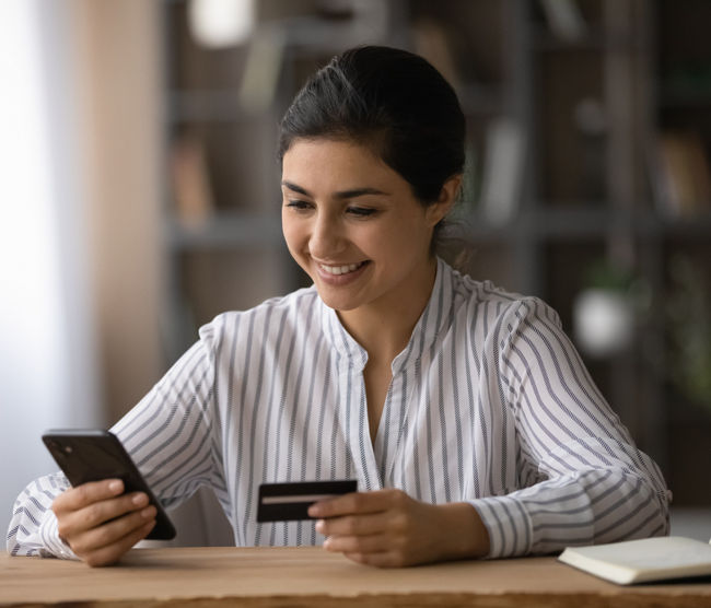 Confident ethnic female student pay tuition fee remotely using mobile phone bank app. Smiling young indian lady sit at office table use credit card to make online money transfer buy goods at internet