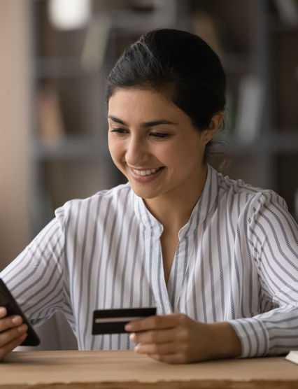 Confident ethnic female student pay tuition fee remotely using mobile phone bank app. Smiling young indian lady sit at office table use credit card to make online money transfer buy goods at internet
