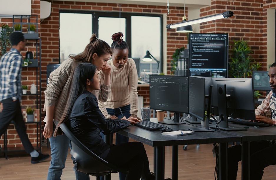 Mixed team of software engineers brainstorming ideas for new code library in front of computer screens compiling algorithms. Diverse app developers collaborating on group project in it agency office.