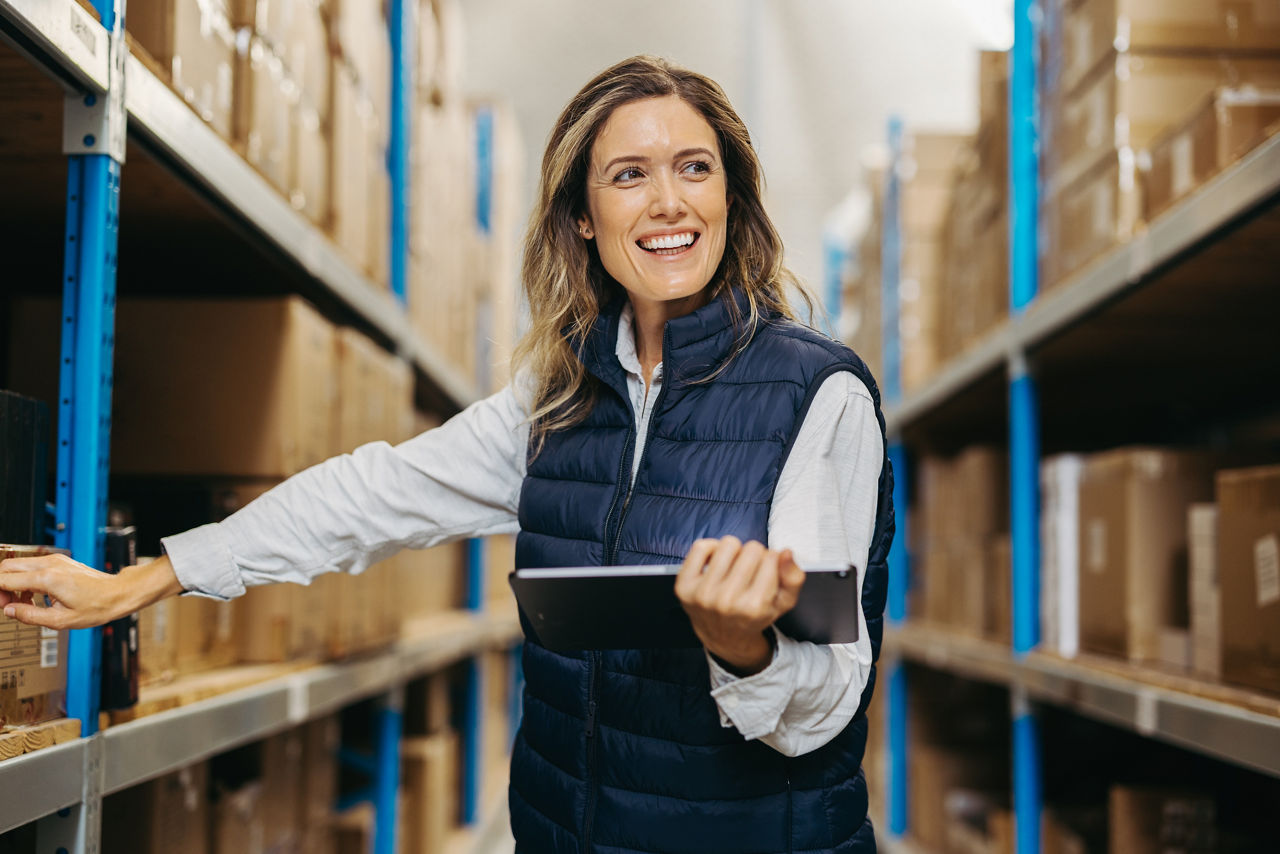 Cheerful warehouse manager smiling while standing with a digital tablet. Happy young woman using smart warehousing technology in a distribution centre.