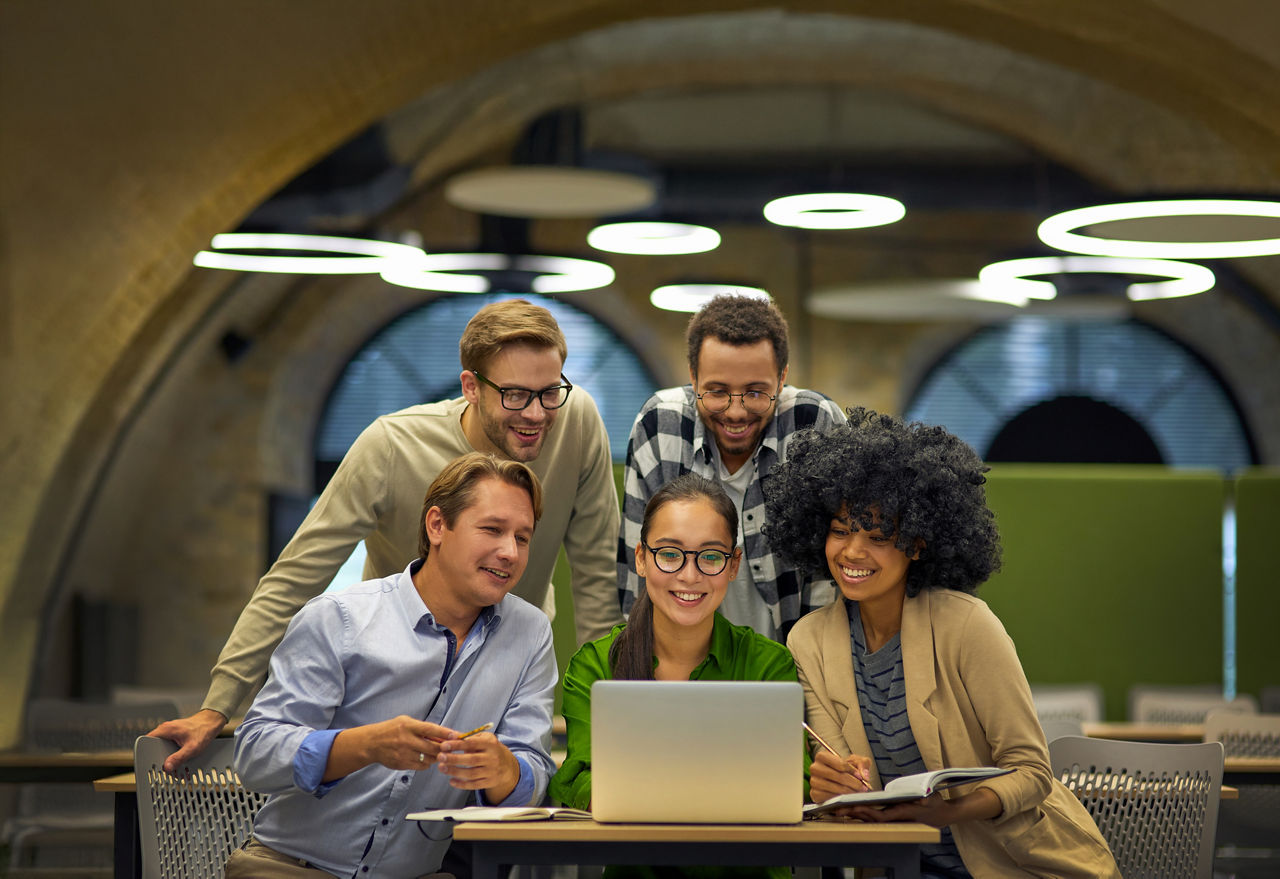 Group of young multiracial business people sitting at desk in the modern coworking space, looking at laptop screen and smiling, working together on a new project