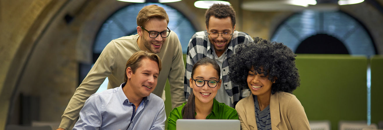 Group of young multiracial business people sitting at desk in the modern coworking space, looking at laptop screen and smiling, working together on a new project