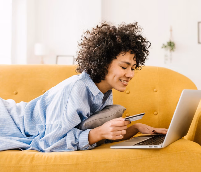Smiling woman holding credit card using laptop lying on sofa in living room at home