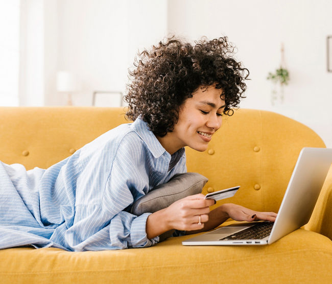 Smiling woman holding credit card using laptop lying on sofa in living room at home