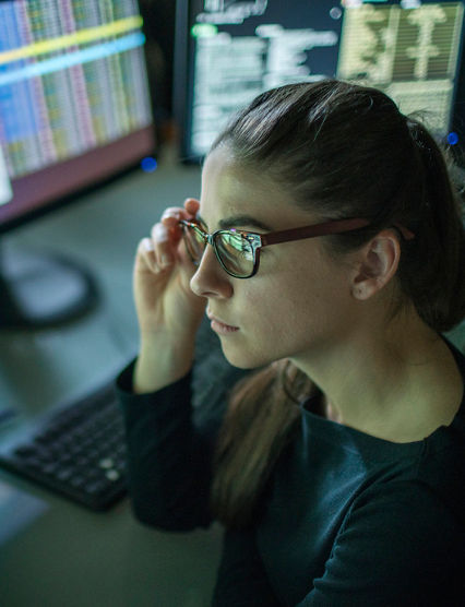 A young woman is seated at a desk surrounded by monitors displaying data, she is contemplating in this dark, moody office.