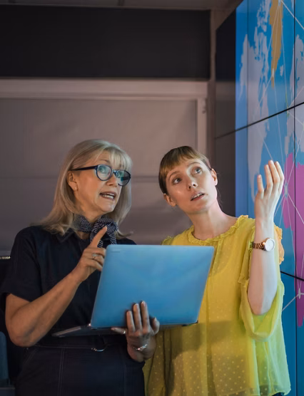 An experienced woman mentors a female colleague, the mature woman is holding a laptop as they debate data from an interactive display; they are both wearing smart casual clothing.