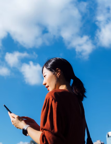 Low angle portrait of young Asian woman using smartphone against beautiful blue sky with cloudscapes, enjoying sunlight outdoors. Lifestyle and technology