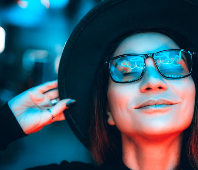 Portrait of a beautiful hipster woman in glasses and hat, she is dancing and dreaming with her eyes closed, enjoying the party night in illuminated neon lights of blue and red. The woman is smiling, reflections in her glasses. Space for text copy