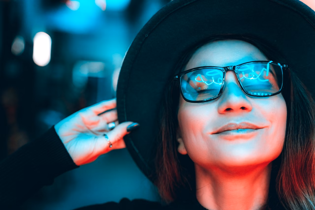 Portrait of a beautiful hipster woman in glasses and hat, she is dancing and dreaming with her eyes closed, enjoying the party night in illuminated neon lights of blue and red. The woman is smiling, reflections in her glasses. Space for text copy