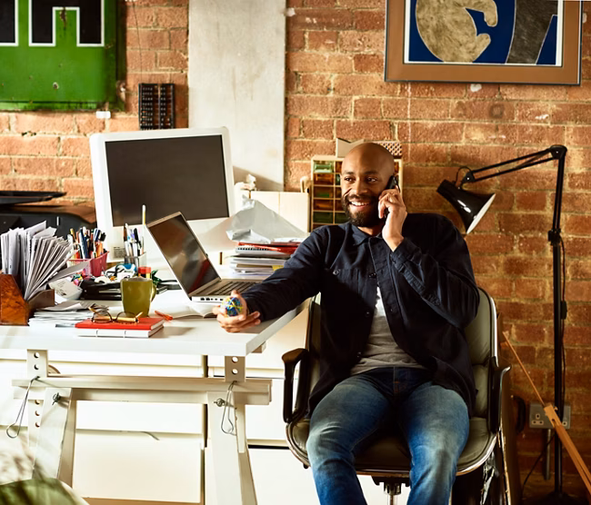 African American small business owner in his 30s talking on smartphone, holding elastic band ball and looking away, confident creative professional in modern studio