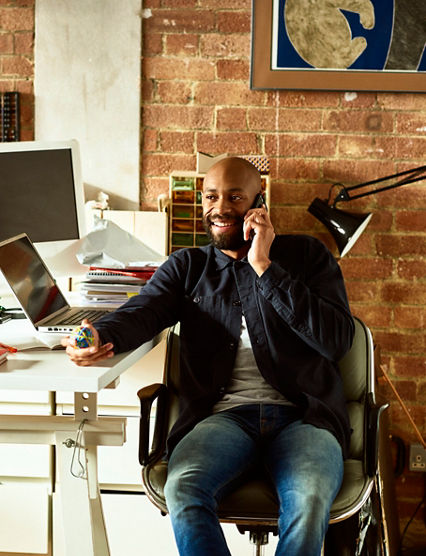 African American small business owner in his 30s talking on smartphone, holding elastic band ball and looking away, confident creative professional in modern studio