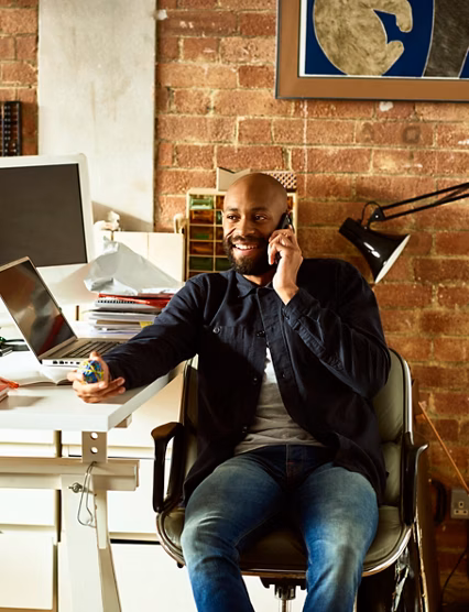 African American small business owner in his 30s talking on smartphone, holding elastic band ball and looking away, confident creative professional in modern studio
