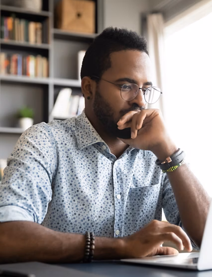 Focused concentrated young african businessman sit at desk look at laptop, serious afro american male professional analyst working online on computer data watch webinar thinking of problem solution