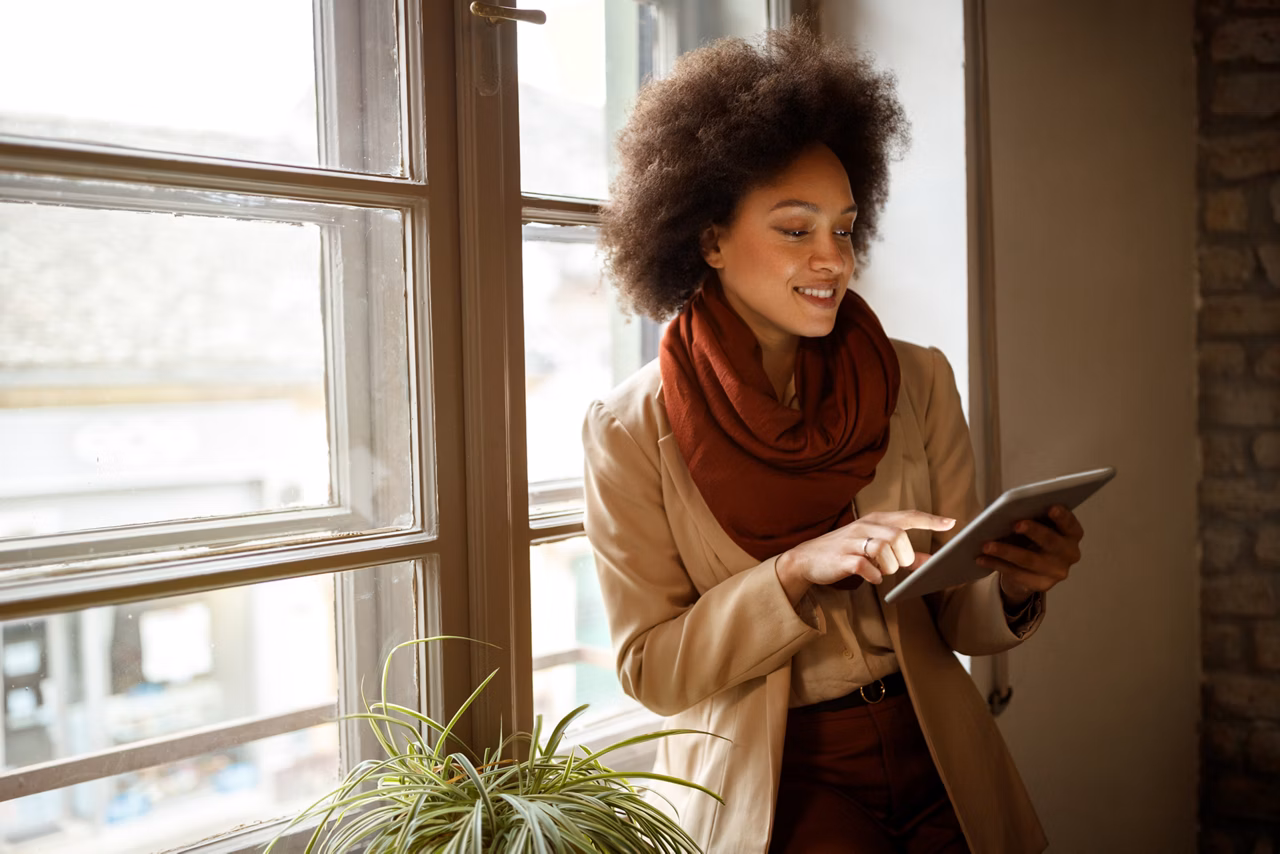 Young Afro-American woman near window on workplace taking information from ipad 