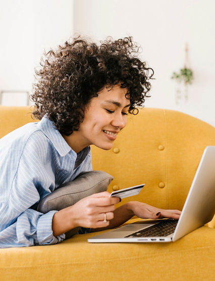 Smiling woman holding credit card using laptop lying on sofa in living room at home