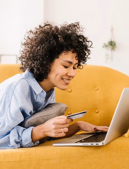 Smiling woman holding credit card using laptop lying on sofa in living room at home