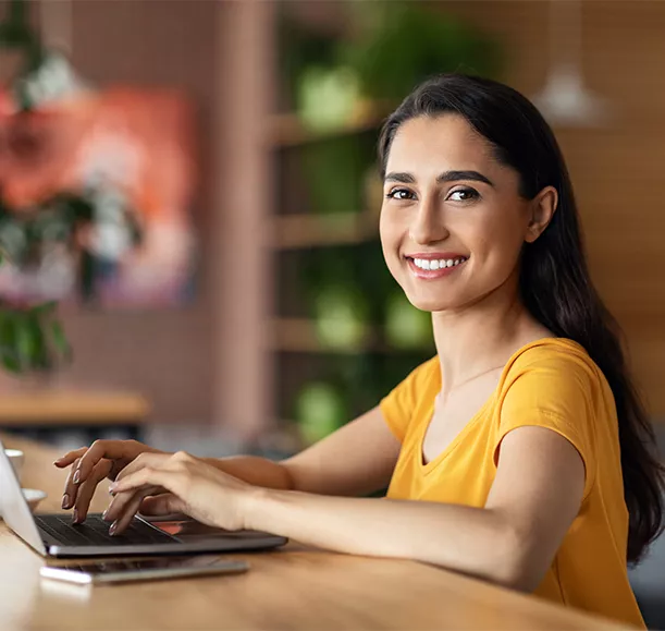 Smiling pretty young arab woman using laptop at cafe, working online, empty space. Side view of cheerful lady freelancer typing on laptop, sending email or chatting with clients, drinking tea
