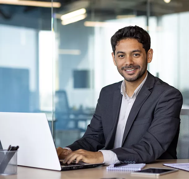 Portrait of young arab businessman, man smiling and looking at camera while sitting inside office, boss in business suit at workplace using laptop.