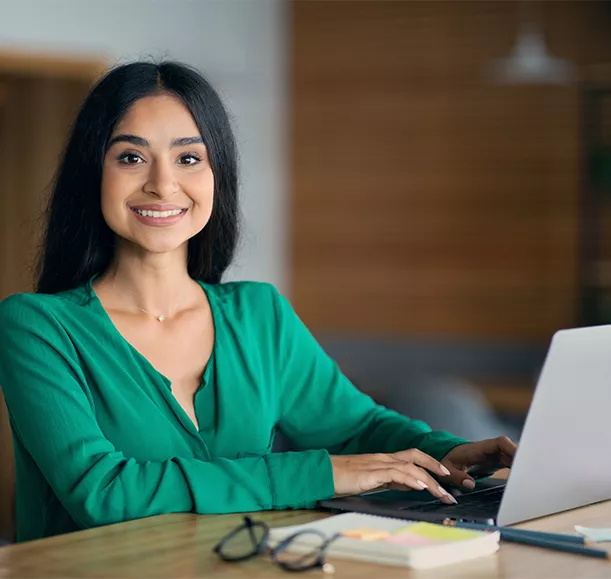 Attractive long-haired indian lady independent contractor working at cafe, sitting at table in front of laptop, typing on keyboard, taking notes, drinking tea, looking at camera, smiling, copy space