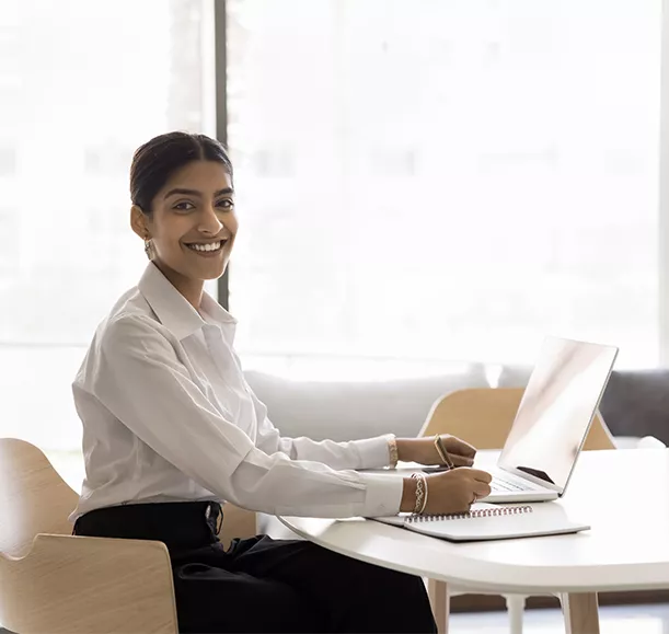 Happy pretty Indian businesswoman office indoor portrait. Young manager, business professional woman working at laptop, sitting at large table, writing learning notes, looking at camera, smiling