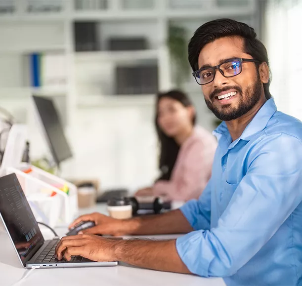 Portrait of an Indian man smiling while working with a laptop at the office and looking at the camera.
