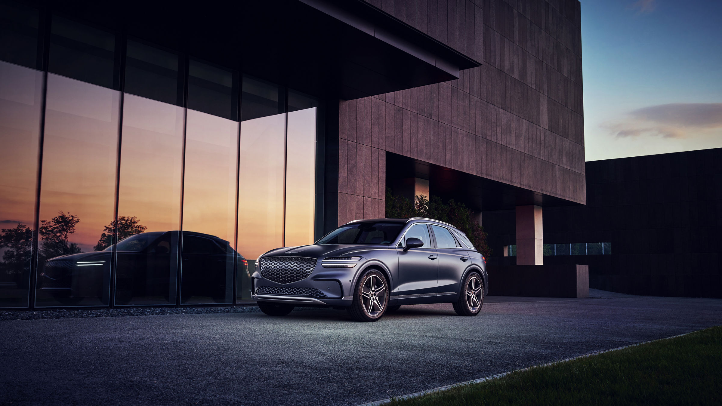 The Ceres Blue Electrified GV70 appears stationary in front of a modern-looking building. The angle provides a view of the vehicle's front and side.