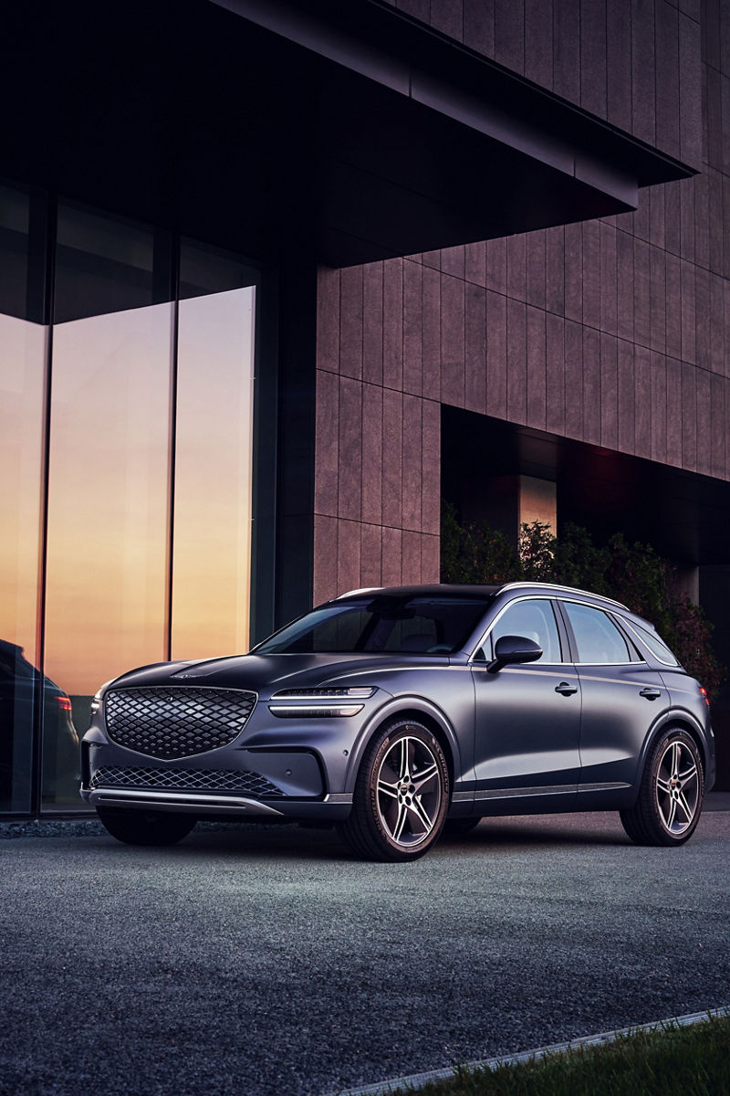 The Ceres Blue Electrified GV70 appears stationary in front of a modern-looking building. The angle provides a view of the vehicle's front and side.