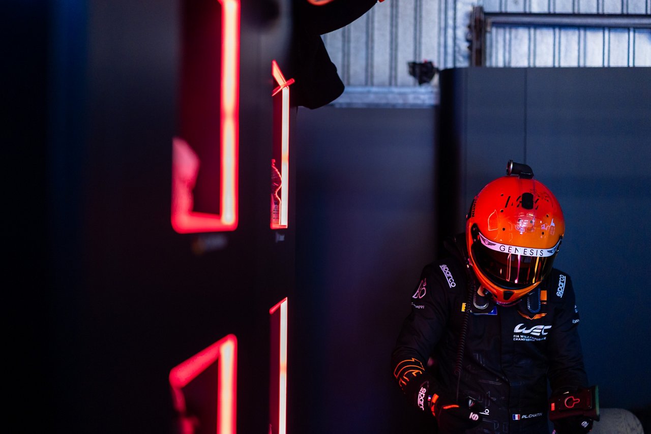 Race crew member in full gear and orange Genesis helmet standing in a dim pit area illuminated by red neon lights.