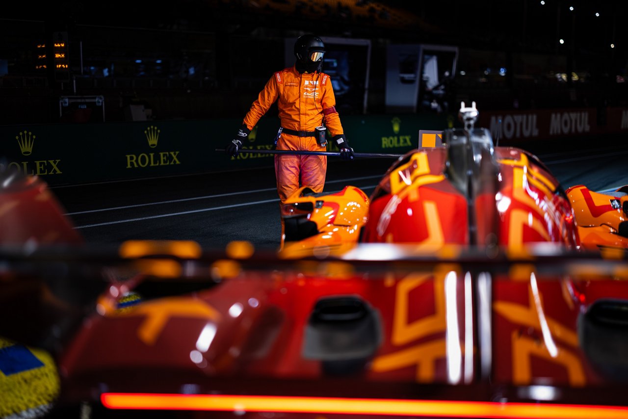 Race marshal in orange suit standing in the pit lane holding a pole as a Genesis GRM-001 waits during a night stop.