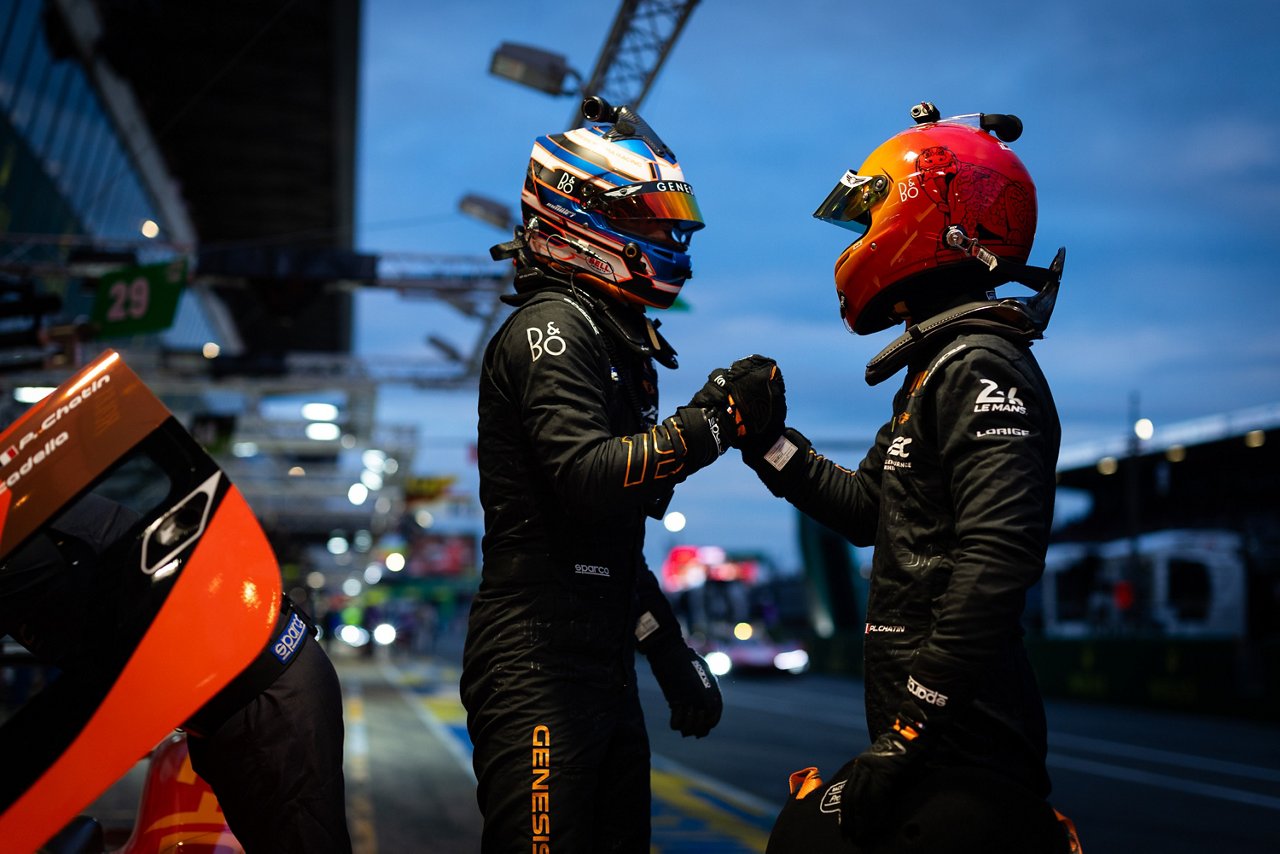 Two Genesis crew members in black suits fist bump in the pit lane beside an orange getaway car at night.