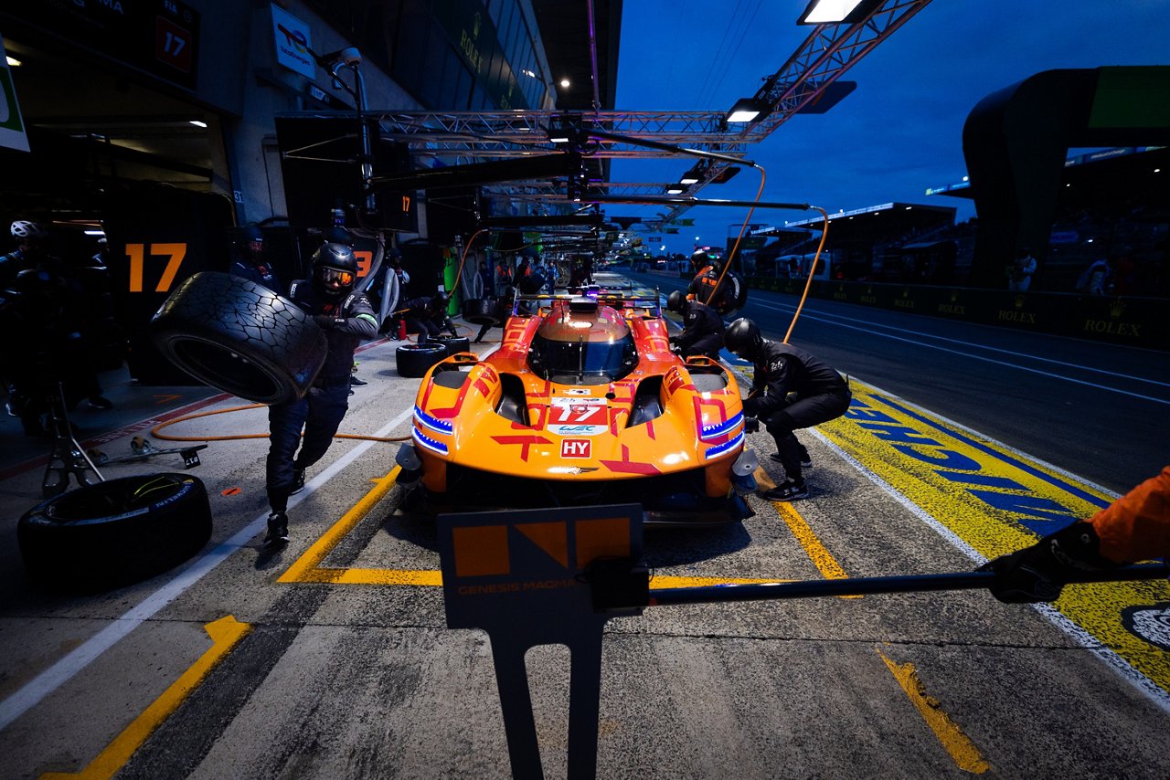 Pit crew servicing the Genesis GRM-001 during a nighttime pit stop, with mechanics changing tires under bright lights.