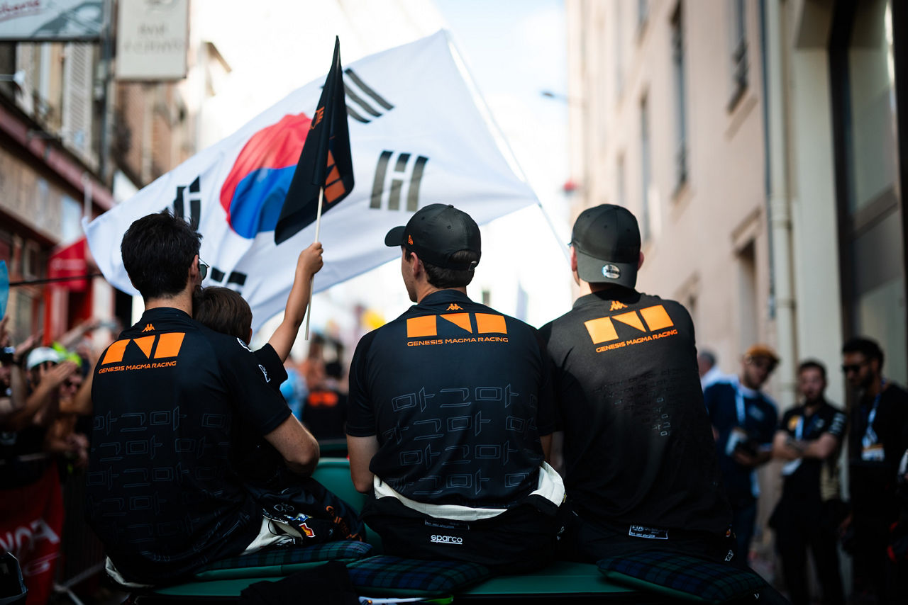 Three Genesis pilots in black “Genesis Magma Racing” shirts ride through a crowded street while holding a South Korean flag.