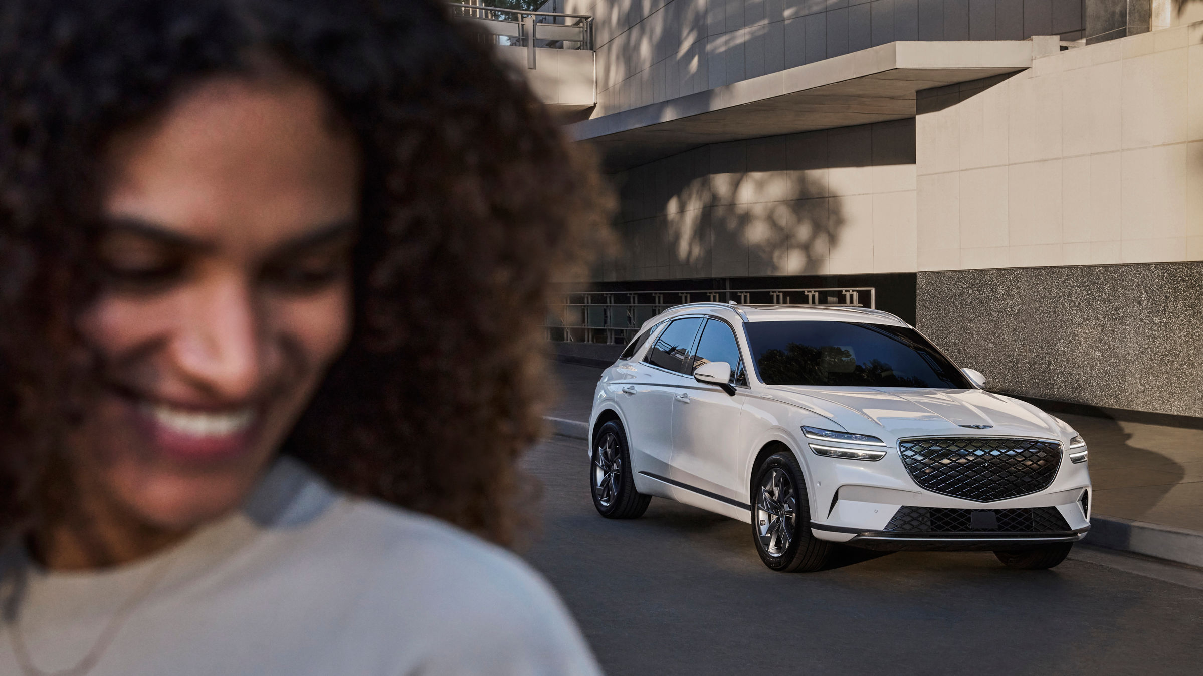 A white Genesis eGV70 parks in front of a house, a smiling woman in the foreground