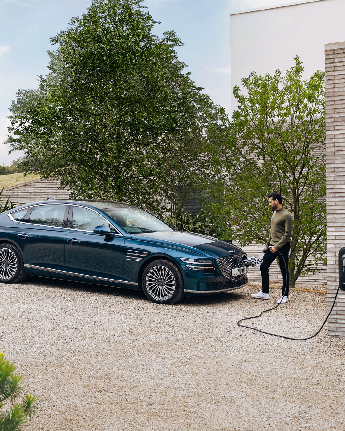 A person is charging a blue Genesis Electrified G80 in the driveway of a house