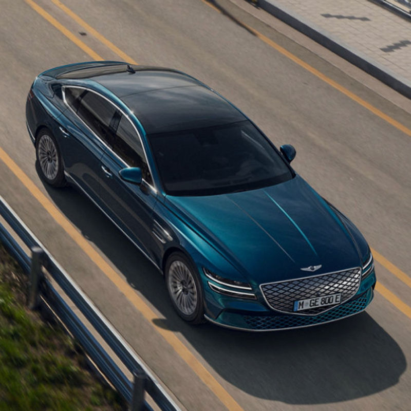 Electrified Genesis G80 in blue driving on a bridge road near the water, top-side view.