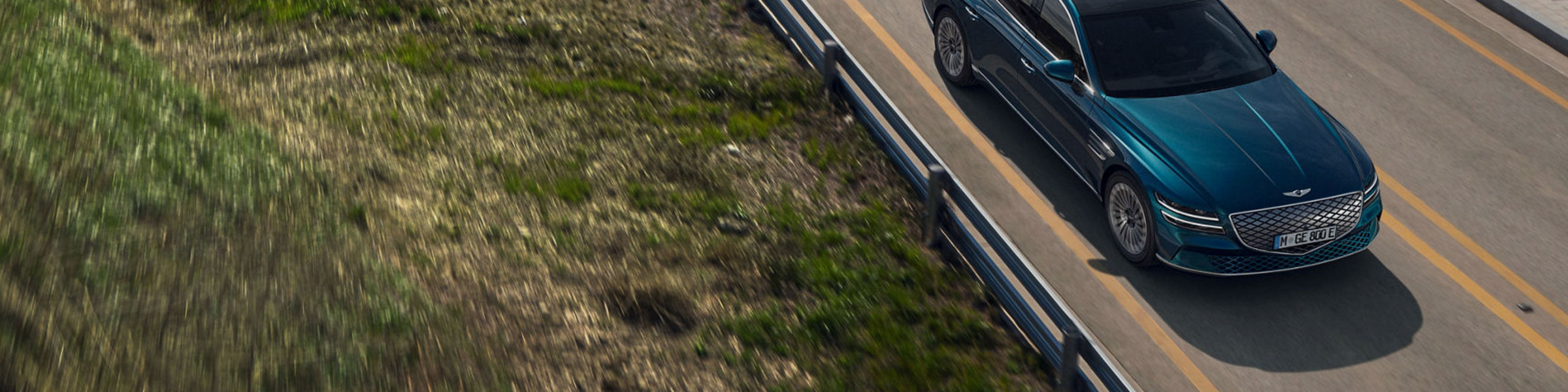 Electrified Genesis G80 in blue driving on a bridge road near the water, top-side view.