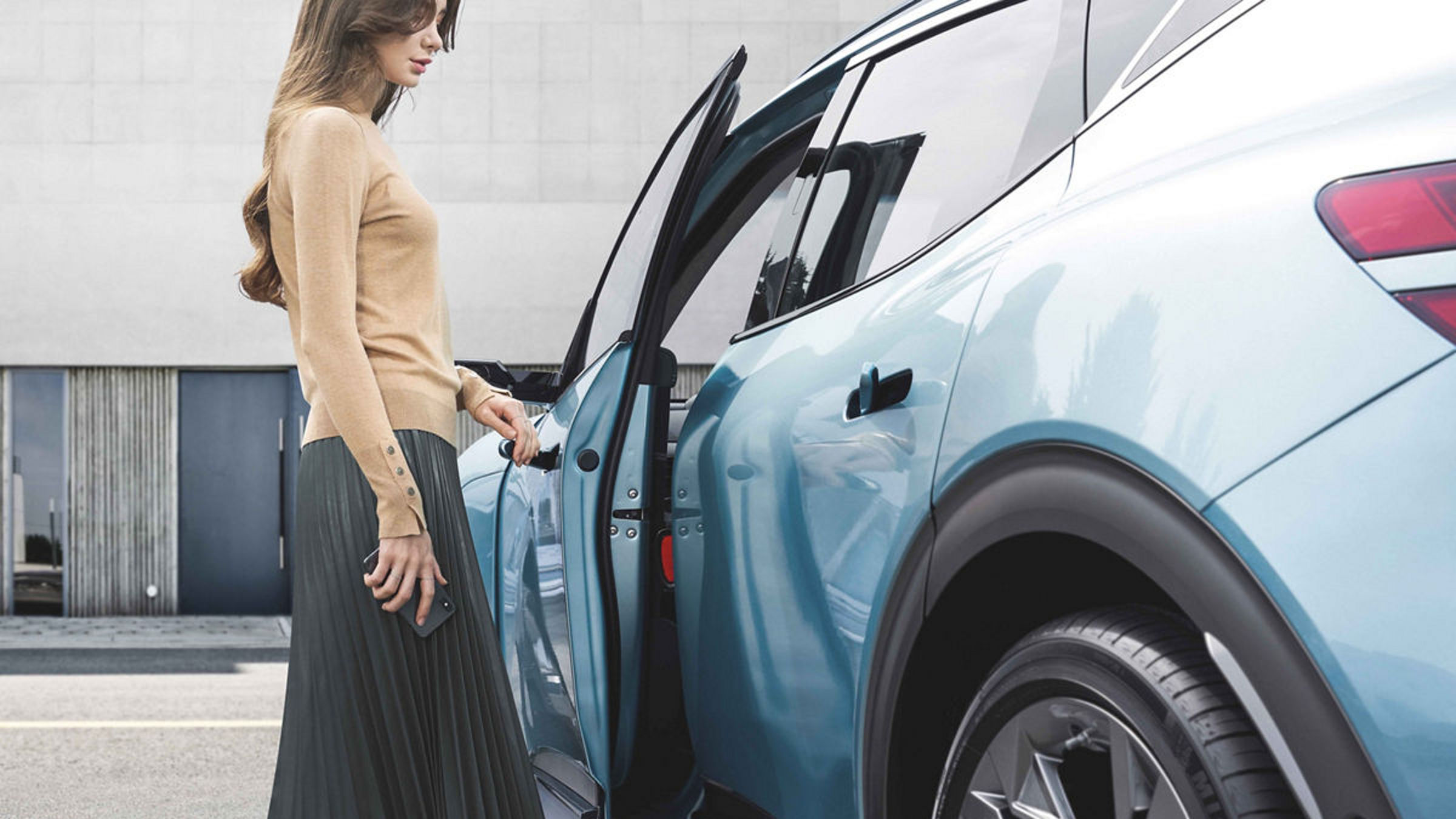 Side view of a woman opening the door of a light blue Genesis car, parked in front of a modern building.