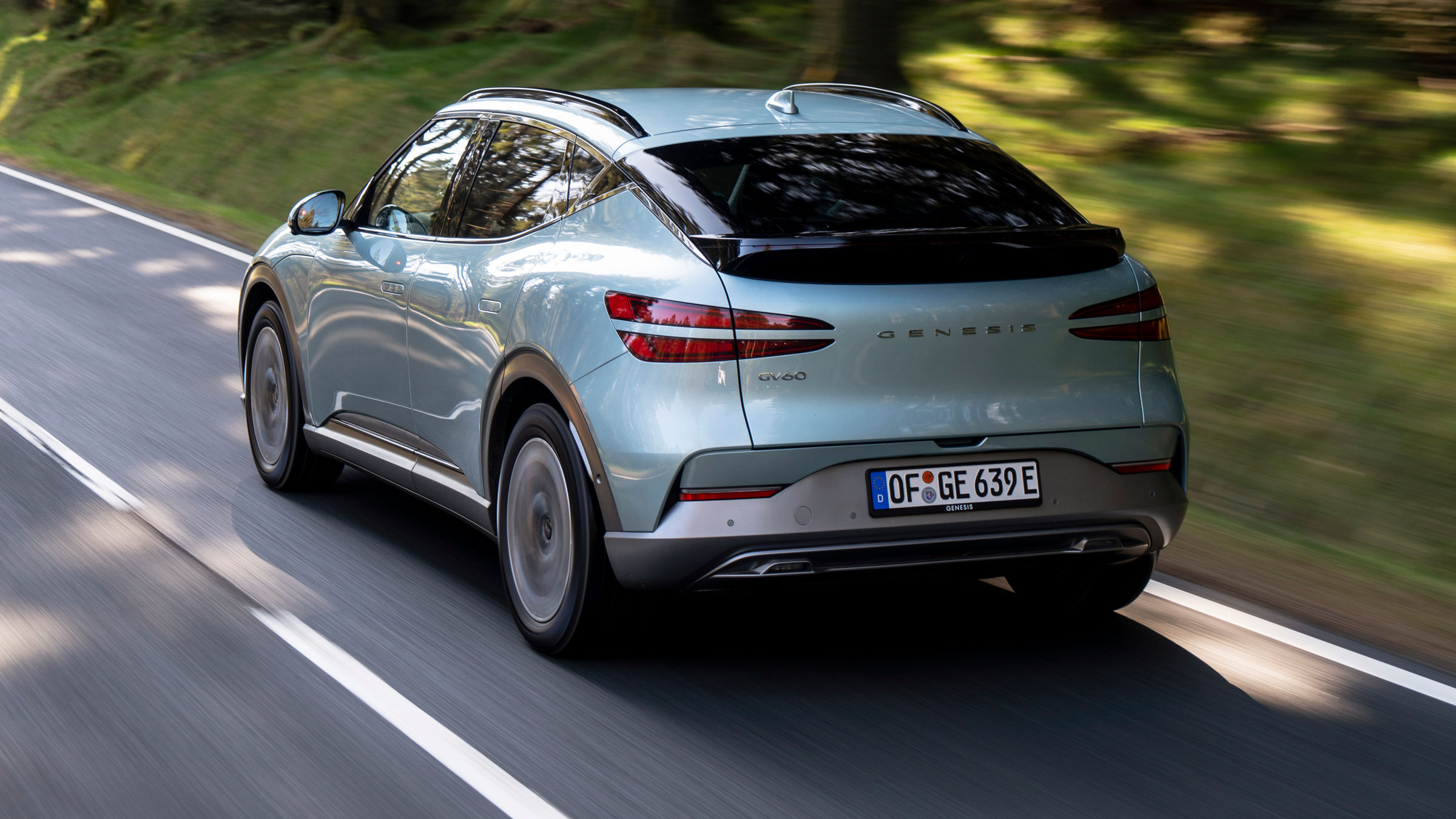 Mint-coloured Genesis GV60 driving through the forest on a highway - side back view