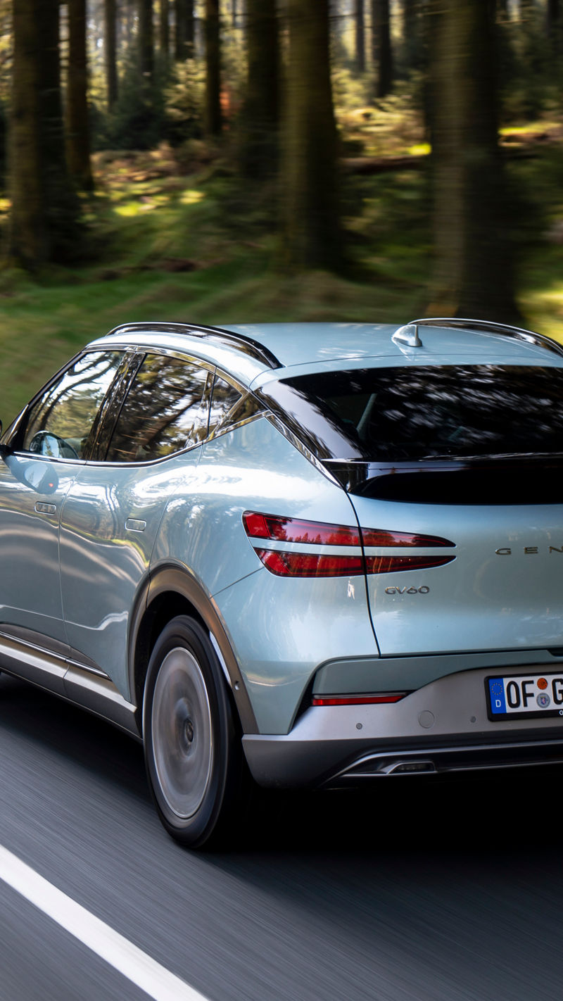 Mint-coloured Genesis GV60 driving through the forest on a highway - side back view