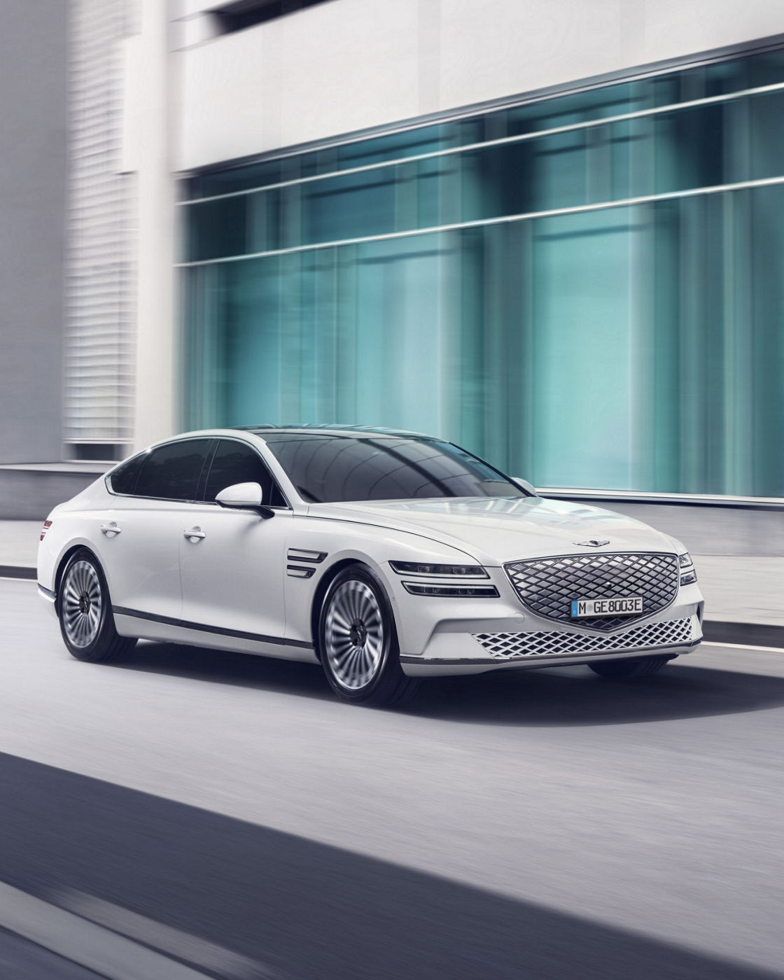  A white Electrified Genesis G80 luxury sedan driving on a city street with modern glass buildings in the background.