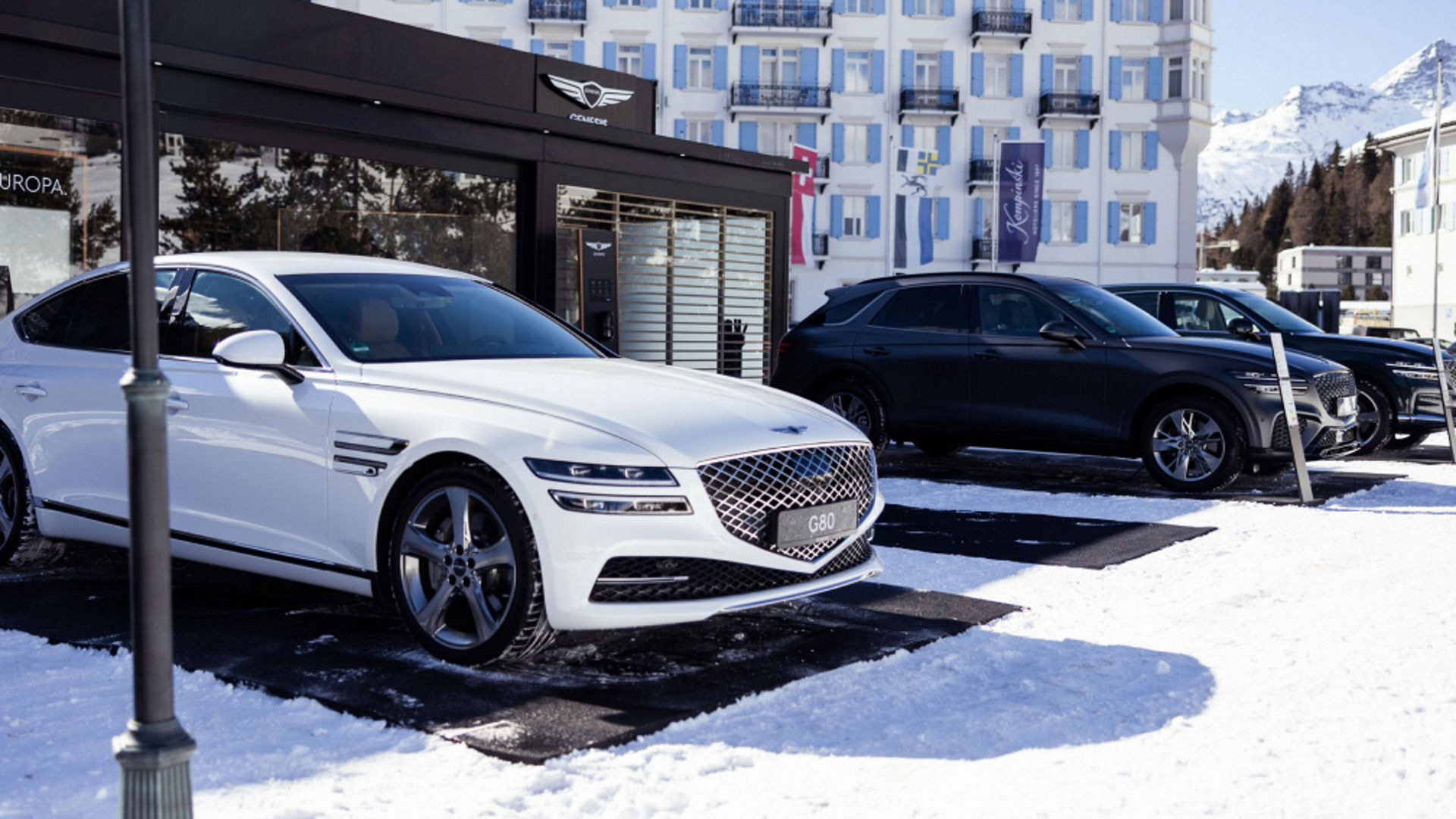 A white Electrified G80 parked on a snowy lot alongside dark SUVs, with a luxury building and mountain peaks in the background.